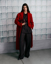 Mannequin devant un mur en briques de verre, portant un long manteau rouge sur un tailleur sombre composé d’un blazer ceinturé et d’un pantalon large. Elle porte des chaussures noires à bout pointu et tient sous le bras un sac bordeaux orné de clous dorés et du logo Love Birds avec les oiseaux emblématiques de PINKO. | Pinko