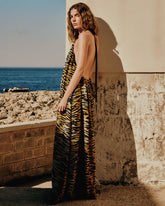A woman wearing a backless tiger-print maxi dress poses against a sun-lit stone wall, with rocky coastline and blue sea in the background. | Pinko
