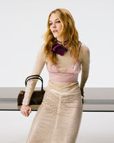 A woman wearing a cream ribbed long-sleeve top with lace inserts, a pink satin bralette, a crystal-embellished midi skirt and a purple flower choker, carrying a dark brown Pinko bowling model bag. | Pinko