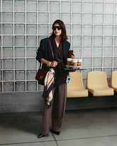 Modella con occhiali da sole, blazer scuro e pantaloni ampi gessati, davanti a una parete in vetrocemento e sedute gialle. Porta a tracolla una borsa bordeaux con logo Love Birds e foulard annodato e regge un vassoio con bicchieri da asporto e un sacchetto di carta. | Pinko