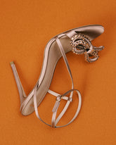 High-heeled sandal with ankle strap in a nude satin finish, embellished with a crystal decorative detail on the front, set against an orange background. | Pinko
