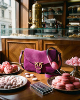 Borsa a tracolla in suede fucsia con logo Pinko e cuffie rosa, posizionate su un tavolo di un caffè italiano circondato da macaron, meringhe e pasticcini rosa. | Pinko