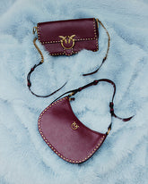 Due borse bordeaux con borchie dorate e logo Love Birds con gli iconici uccellini PINKO, adagiate su una superficie azzurra dall’effetto pelliccia. In alto, un modello rettangolare con patta e tracolla con inserti a catena; in basso, un modello a mezzaluna con cinturino sottile. | Pinko