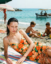 Eine Frau liegt auf einem gestreiften Strandtuch und trägt ein orangefarbenes Blumen-Minikleid mit Fransenddetail, umgeben von einer lebhaften Strandszene mit spielenden Kindern im Sand und dem Meer im Hintergrund. | Pinko