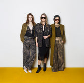 Three women standing on a yellow and white background, wearing khaki green and black total looks: camouflower-print trousers, a black dress with a printed jacket and sunglasses, and an olive coat with metallic trousers. | Pinko