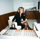 Donna bionda seduta in uno studio radiofonico con insegna al neon "Pinko Radio", indossa un abito nero con dettaglio a stringatura in vita e décolleté rosse, con cuffie e microfono, tiene in mano una borsa nera. | Pinko