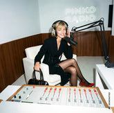 Donna bionda seduta in uno studio radiofonico con insegna al neon "Pinko Radio", indossa un abito nero con dettaglio a stringatura in vita e décolleté rosse, con cuffie e microfono, tiene in mano una borsa nera. | Pinko