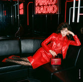 Dark-haired woman reclining on a black leather sofa in a venue with "Pinko Radio" neon signs, wearing a red leather jacket and skirt set, logo-patterned tights and red pumps, holding a small red vanity-case-shaped bag. | Pinko