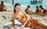 Eine Frau liegt auf einem gestreiften Strandtuch und trägt ein orangefarbenes Blumen-Minikleid mit Fransenddetail, umgeben von einer lebhaften Strandszene mit spielenden Kindern im Sand und dem Meer im Hintergrund. | Pinko