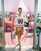 Eine Frau in einem weißen Pinko-Football-T-Shirt „Bold Soul 90", einem orangefarbenen Blumen-Minirock mit Fransen und schwarzen Riemchen-Absatzsandalen, mit einer hellblauen Pinko-Tasche, die durch einen rosafarbenen Perlenvorhang geht, mit einer Retro-Strandszene im Hintergrund. | Pinko