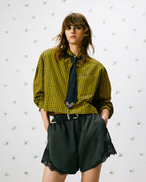 Modella in posa frontale con camicia cropped a quadri gialli e neri, cravatta nera e shorts ampi con dettagli in pizzo nero, su sfondo chiaro con micro motivo floreale. | Pinko