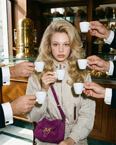 Donna bionda con capelli mossi seduta in un caffè italiano, circondata da mani che le porgono tazzine di caffè espresso, indossa un giubbotto beige e una borsa a tracolla in suede viola con logo Pinko. | Pinko