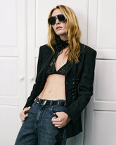 Model mit schwarzem Blazer mit Schnürdetails, schwarzem perforiertem Bralette, High-Waist-Jeans, übergroßer Sonnenbrille und Lederhalstuch vor weißem Wandpaneel-Hintergrund. | Pinko