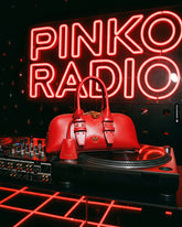 Red handbag with the Pinko logo resting on a turntable next to a DJ mixer, in front of the red "Pinko Radio" neon sign. | Pinko