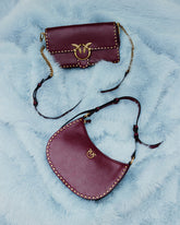 Due borse bordeaux con borchie dorate e logo Love Birds con gli iconici uccellini PINKO, adagiate su una superficie azzurra dall’effetto pelliccia. In alto, un modello rettangolare con patta e tracolla con inserti a catena; in basso, un modello a mezzaluna con cinturino sottile. | Pinko