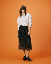 Modella in posa frontale con camicia bianca a righe dal taglio morbido, gonna midi nera semitrasparente con texture luminosa e scarpe stringate nere, su sfondo arancione uniforme. | Pinko