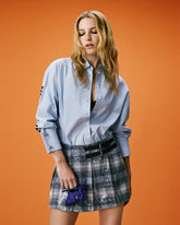 Donna con capelli biondi ondulati su sfondo arancione, indossa una camicia azzurra oversize e una minigonna plissettata a quadri con paillettes e cinturini neri, con una piccola borsa blu elettrico in mano. | Pinko
