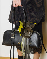 Close-up of a black bowling bag with the Pinko logo, decorated with an olive green and yellow flower-shaped brooch with fringe, held by a woman wearing a camouflower-print dress. | Pinko