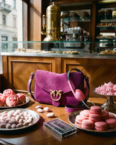 Borsa a tracolla in suede fucsia con logo Pinko e cuffie rosa, posizionate su un tavolo di un caffè italiano circondato da macaron, meringhe e pasticcini rosa. | Pinko