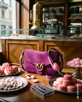 Borsa a tracolla in suede fucsia con logo Pinko e cuffie rosa, posizionate su un tavolo di un caffè italiano circondato da macaron, meringhe e pasticcini rosa. | Pinko