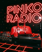 Red handbag with the Pinko logo resting on a turntable next to a DJ mixer, in front of the red "Pinko Radio" neon sign. | Pinko