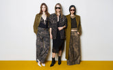Three women standing on a yellow and white background, wearing khaki green and black total looks: camouflower-print trousers, a black dress with a printed jacket and sunglasses, and an olive coat with metallic trousers. | Pinko