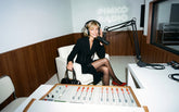 Donna bionda seduta in uno studio radiofonico con insegna al neon "Pinko Radio", indossa un abito nero con dettaglio a stringatura in vita e décolleté rosse, con cuffie e microfono, tiene in mano una borsa nera. | Pinko