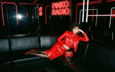 Dark-haired woman reclining on a black leather sofa in a venue with "Pinko Radio" neon signs, wearing a red leather jacket and skirt set, logo-patterned tights and red pumps, holding a small red vanity-case-shaped bag. | Pinko