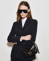 Model wearing oversized black sunglasses, a black suit and a white shirt, standing against a light wall. Her arms are crossed as she holds the strap of a glossy black flap bag with a gold-tone Love Birds logo featuring the iconic PINKO birds. | Pinko